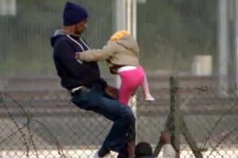A-migrant-straddling-a-fence-with-a-young-child-at-Calais